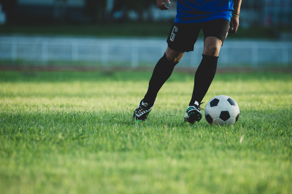 Hráč futbalu driblí loptu na trávniku, oblečený v modrom drese.